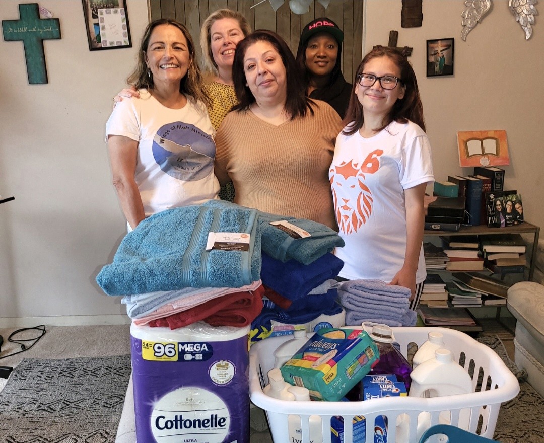 Five women standing behind a table with folded towels, laundry supplies, and a laundry basket in a room with wall decorations.