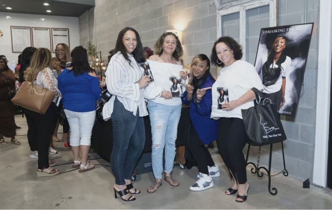 Group of four women holding photographs, standing near a poster of a woman, with people in line in background.
