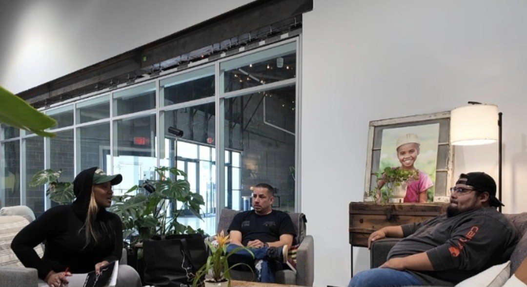Three people sitting in a modern indoor space with large windows, plants, and artwork on the wall.