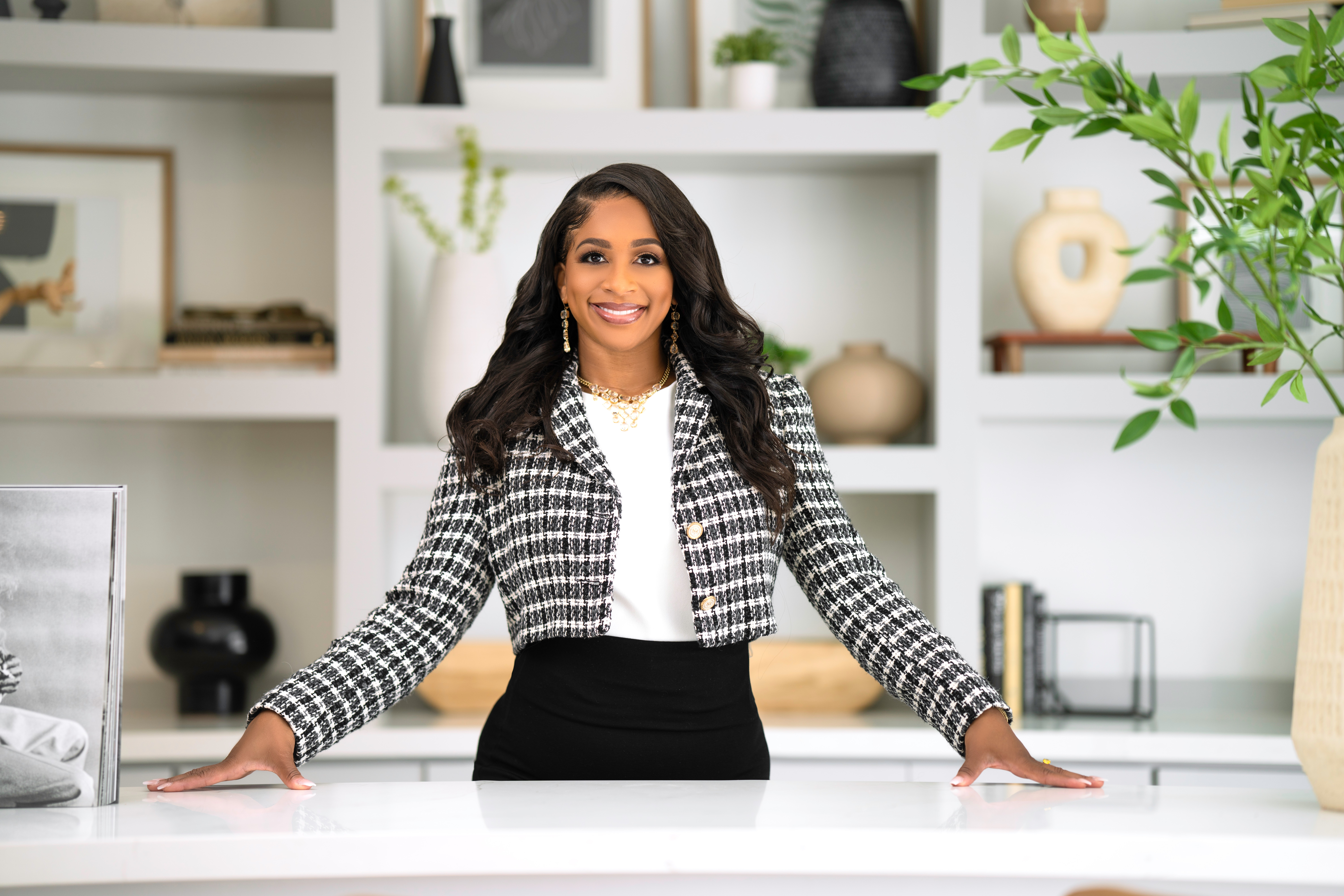 Woman with long dark hair smiling, standing behind a white desk in a modern, decorated office space.