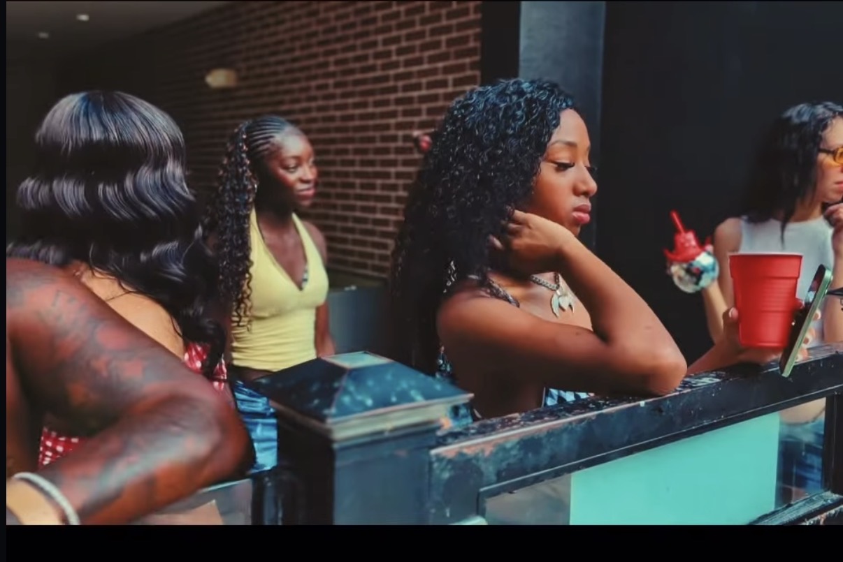 Four women standing at a counter, one with a red cup and a dessert, in front of a brick wall.