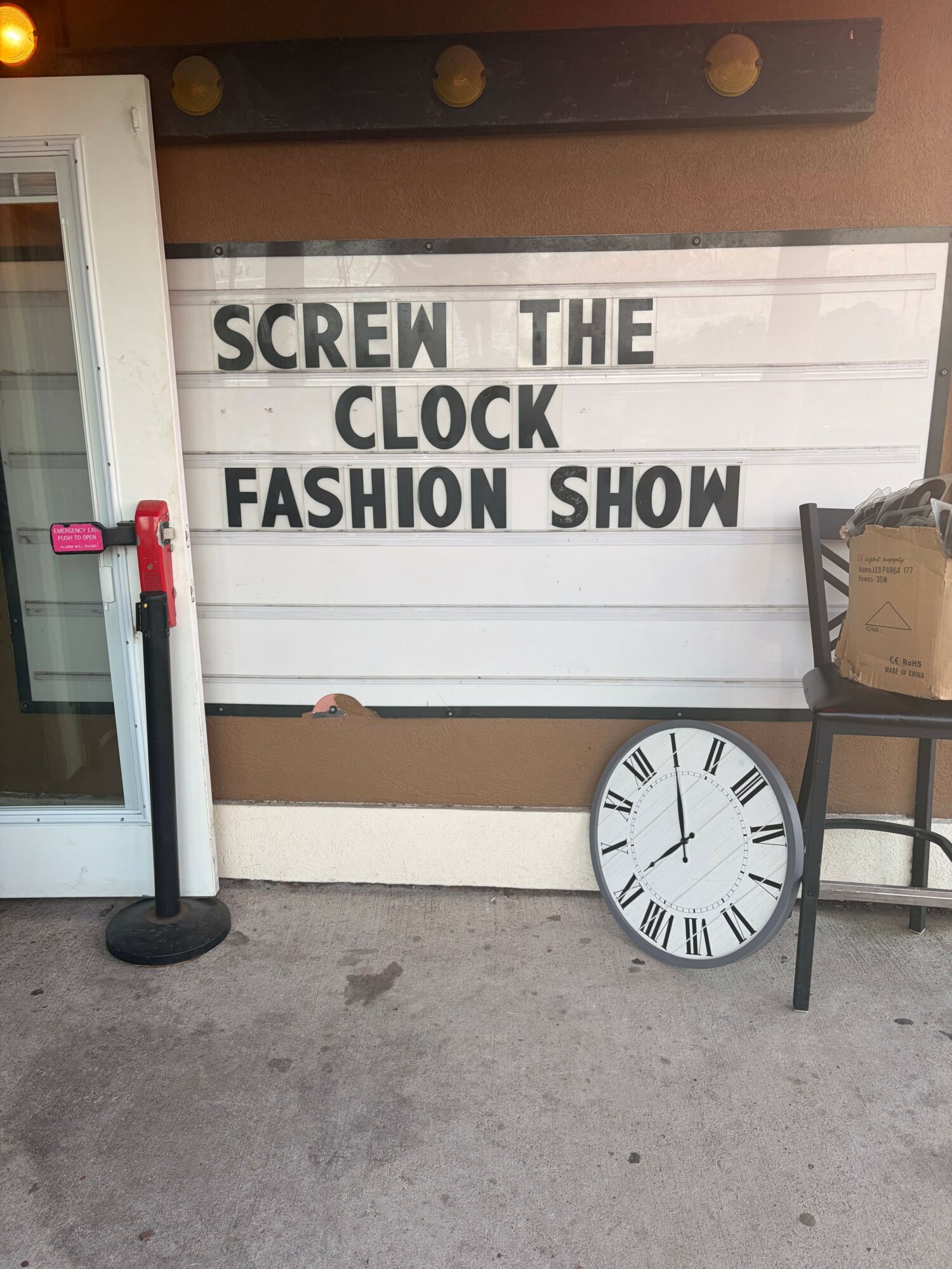 Clock on the ground leaning against a white stand, with a sign behind it reading 'SCREEN THE CLOCK! FASHION SHOW'.