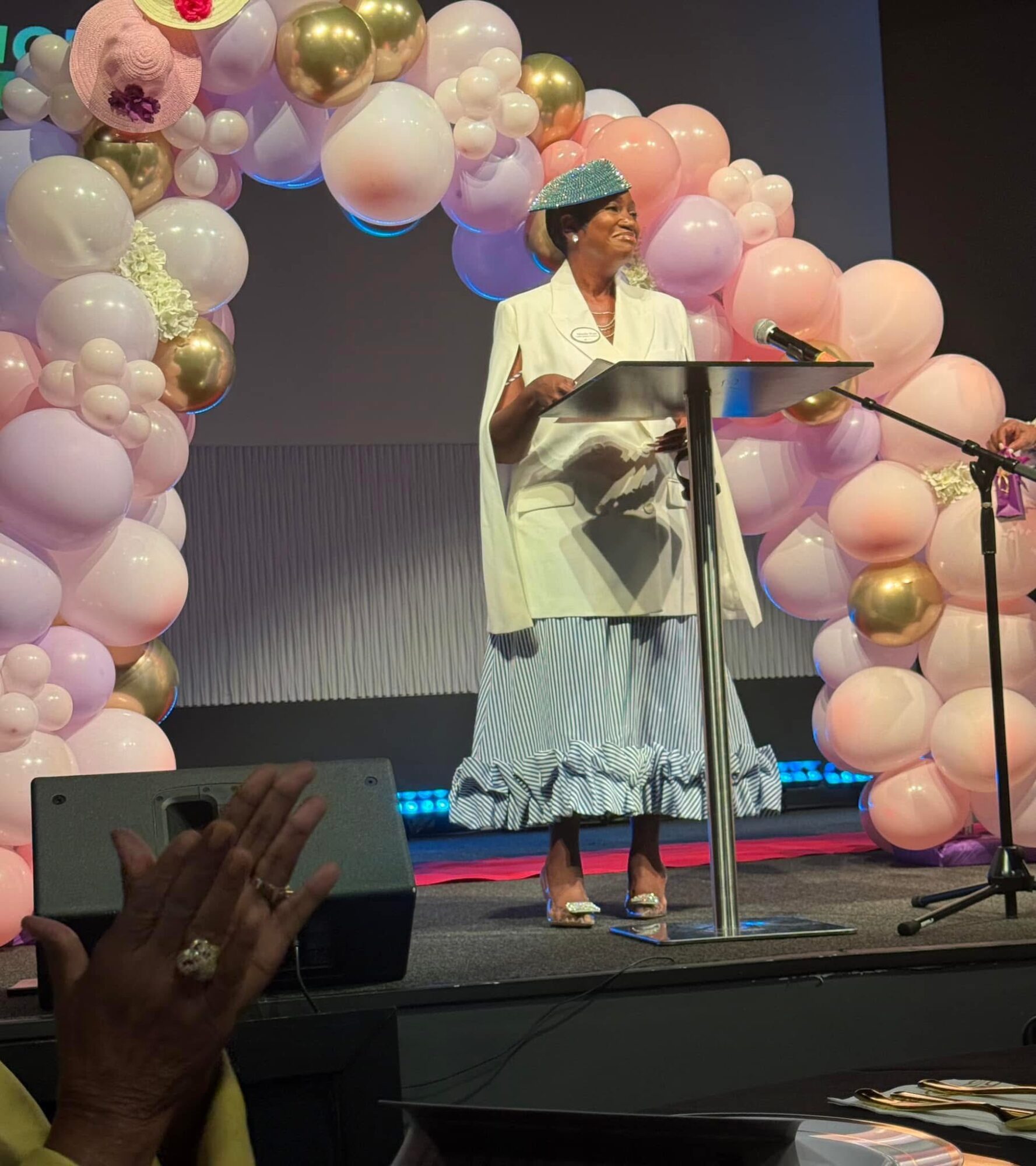 Woman in white dress and headwrap speaking at a podium on stage decorated with pink, white, and gold balloons.