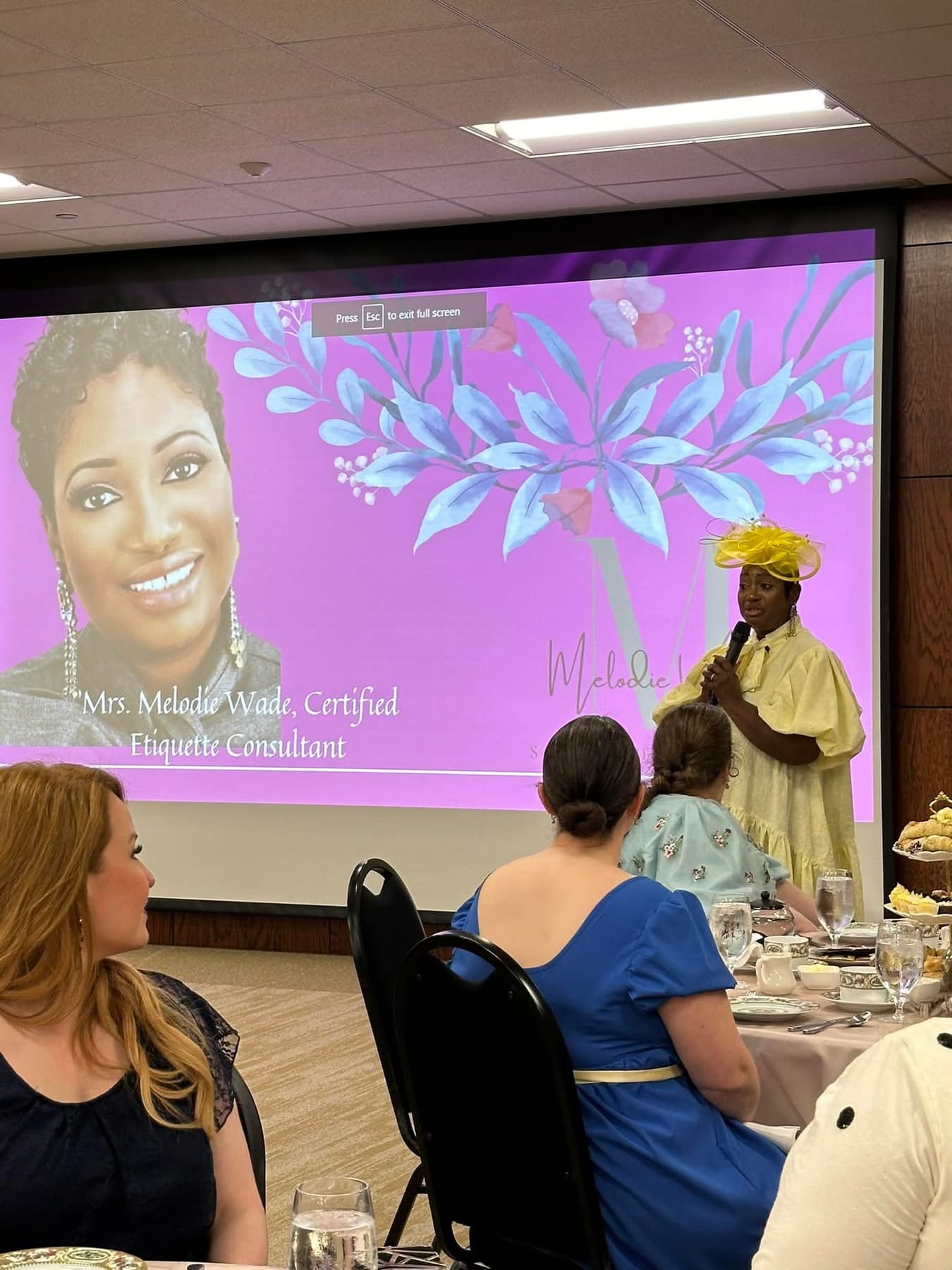 Woman in yellow headscarf speaking to seated audience in a room with a large screen displaying a smiling woman's portrait and purple floral background.