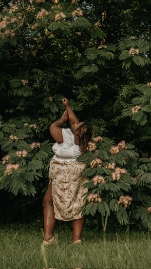 Person standing outdoors among lush green foliage, facing away, with arms raised above head, wearing a white top and patterned skirt.