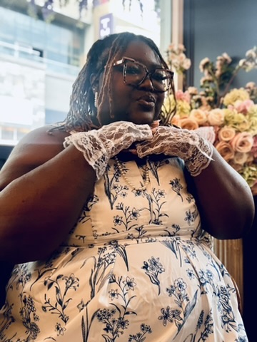Woman with glasses and dreadlocks wearing a floral dress, holding lace gloves near her face, in front of a window with flowers.