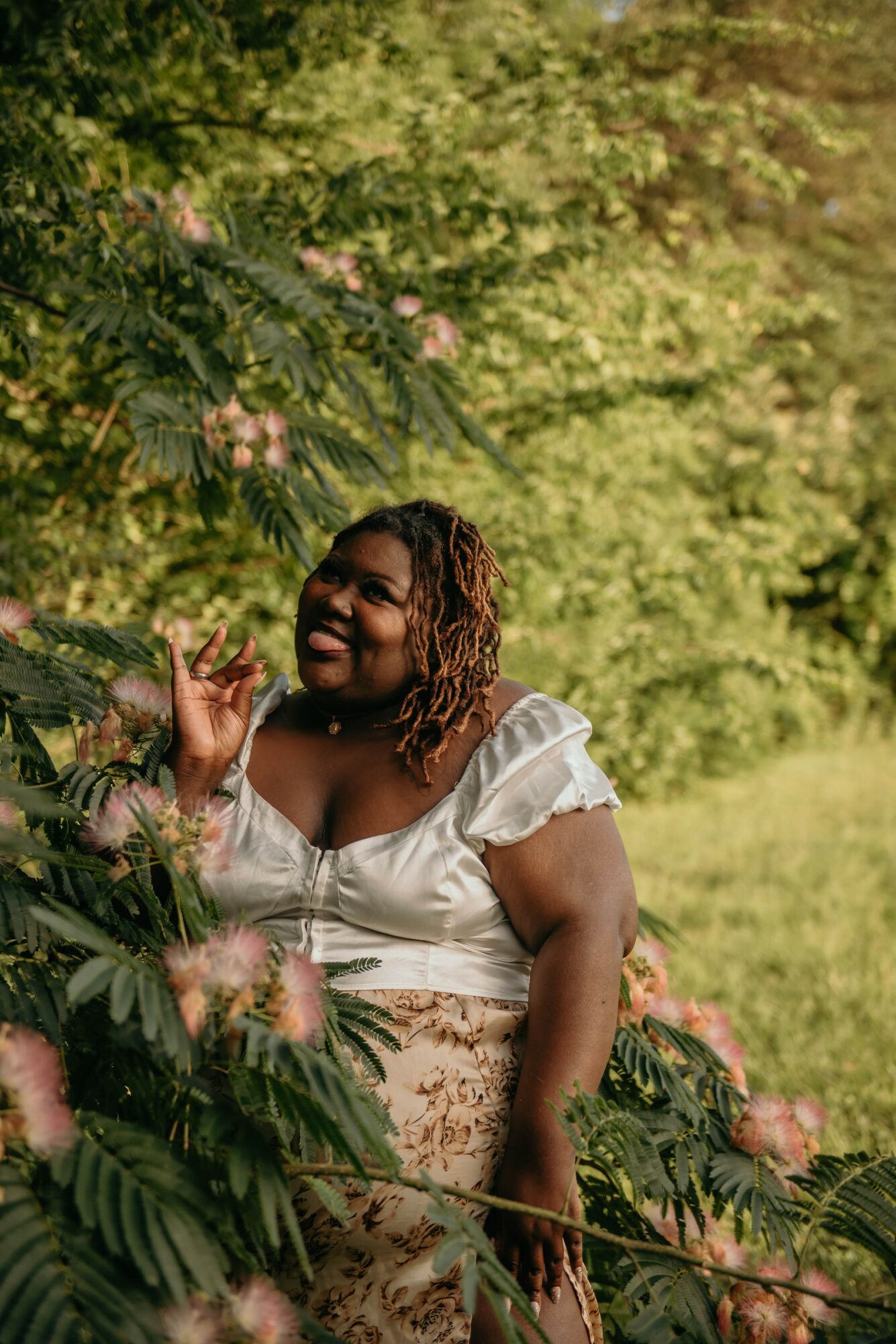 Smiling woman with curly hair standing outdoors among pink flowers and green foliage.