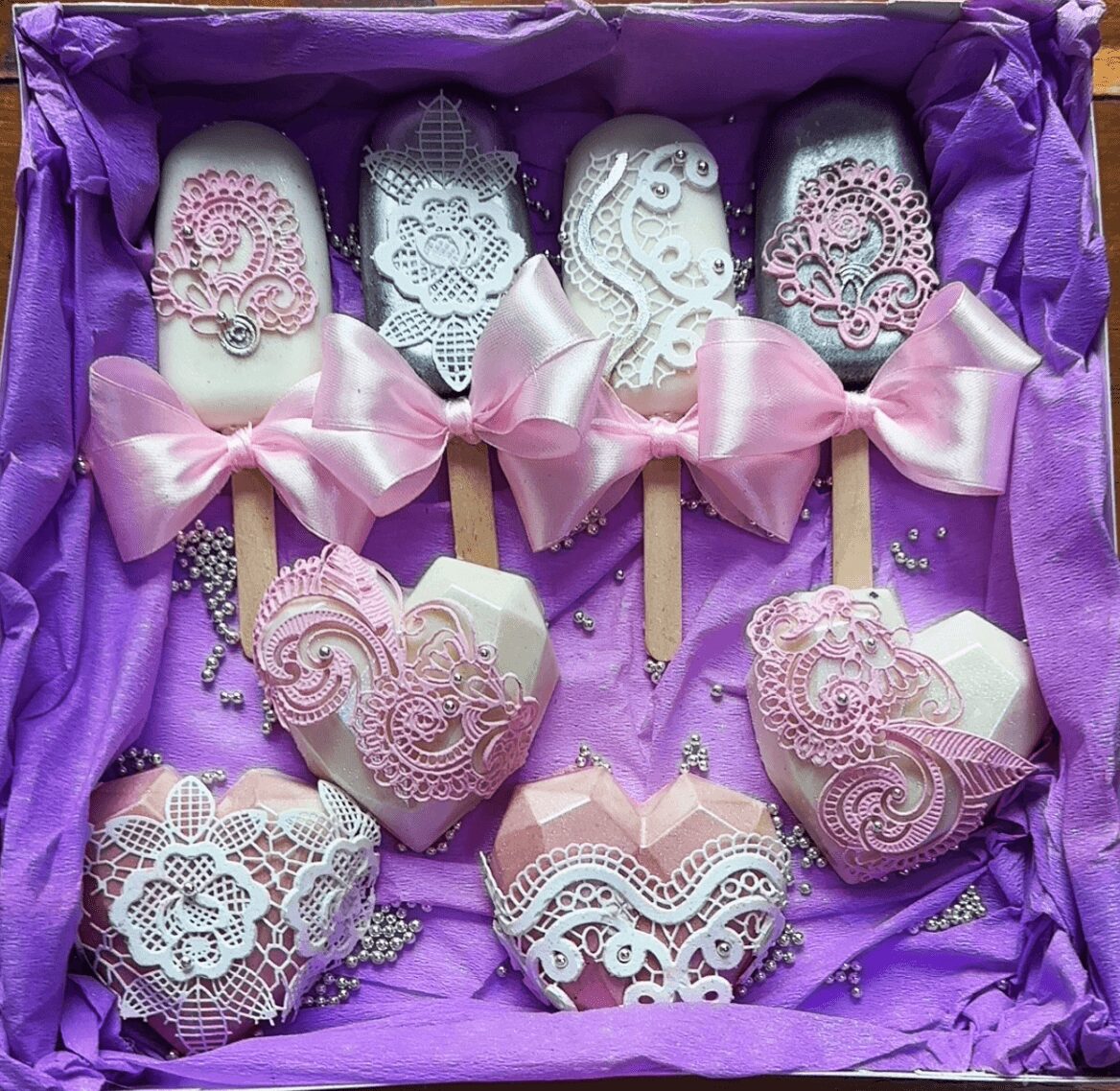 Decorative cookies with lace and floral patterns on sticks, pink bows, in purple tissue paper, with pearl-like beads, for celebration.