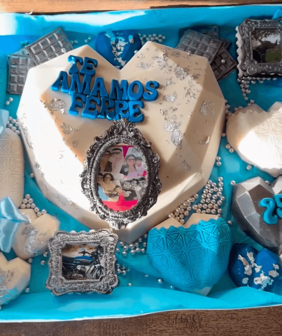 Heart-shaped cake with blue and white decorations, surrounded by small picture frames and chocolates, celebrating a special occasion.