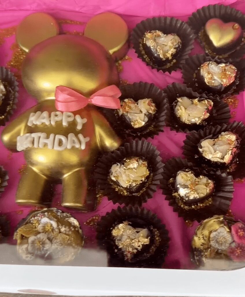 Gold bear-shaped birthday cake with a pink bow, surrounded by chocolates with gold and heart-shaped toppings, on pink background.