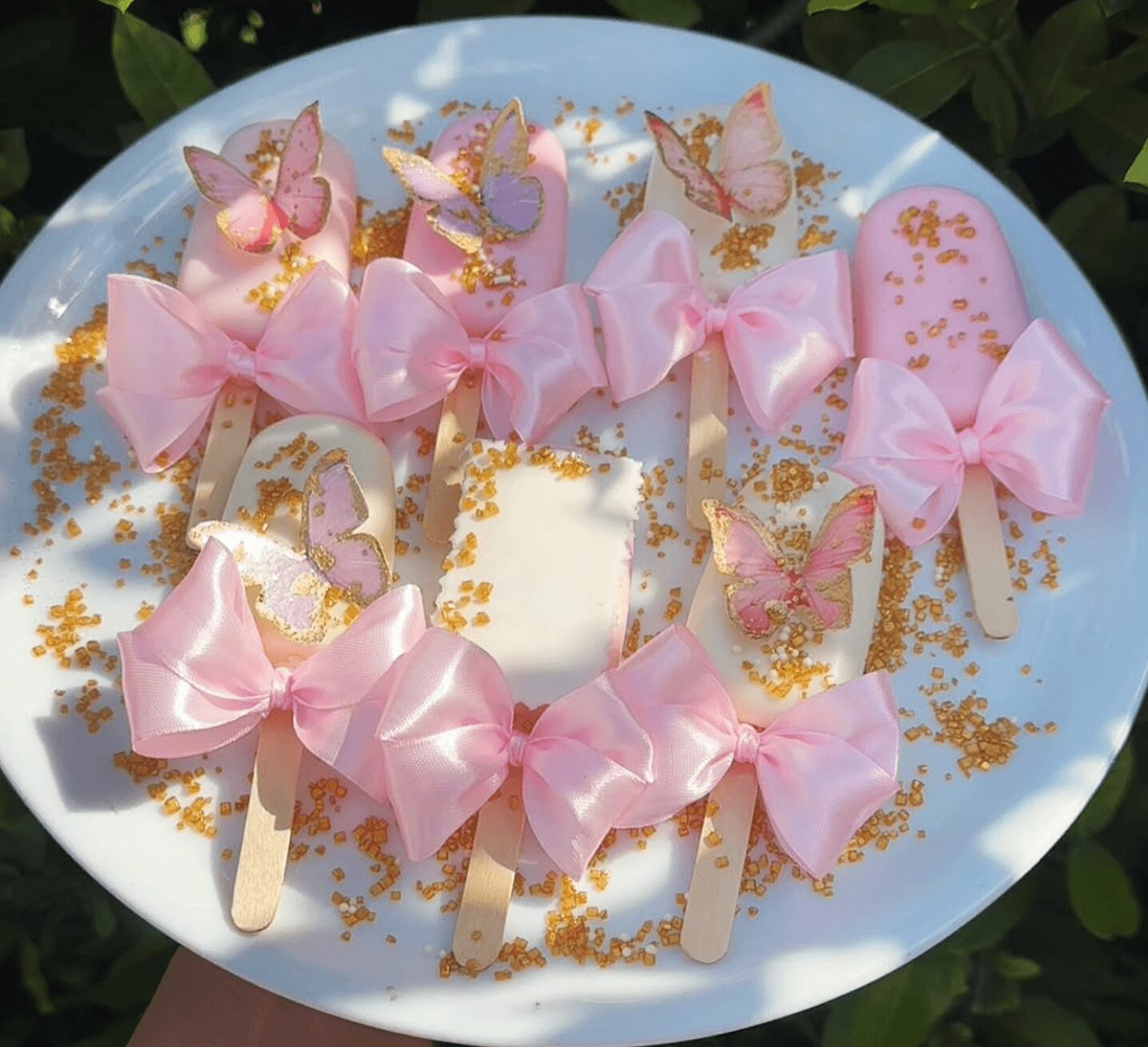 Decorative popsicles with pink bows, butterfly designs, and gold sprinkles on a blue plate.