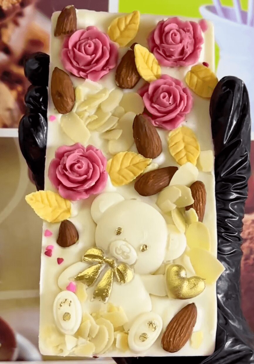 Decorative cake with pink roses, yellow leaves, almonds, and a white teddy bear with a gold bow, on a white surface.