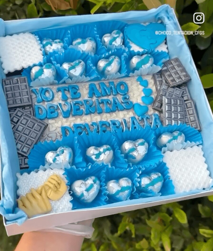 Blue box with chocolates, a message in blue letters, and decorative elements, held outdoors with green foliage in background.