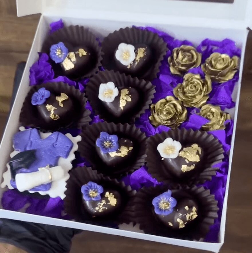 Box of decorated chocolates with purple flowers and gold accents, arranged with purple tissue paper, in a white box.