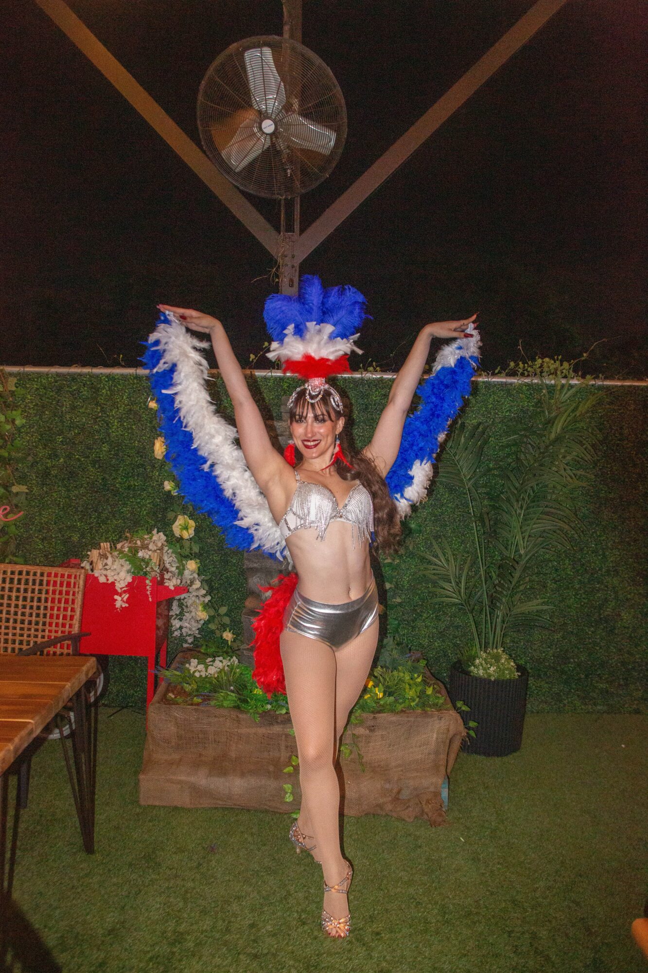 Performer in silver costume with blue and white feathered wings, arms raised, standing outdoors at night.
