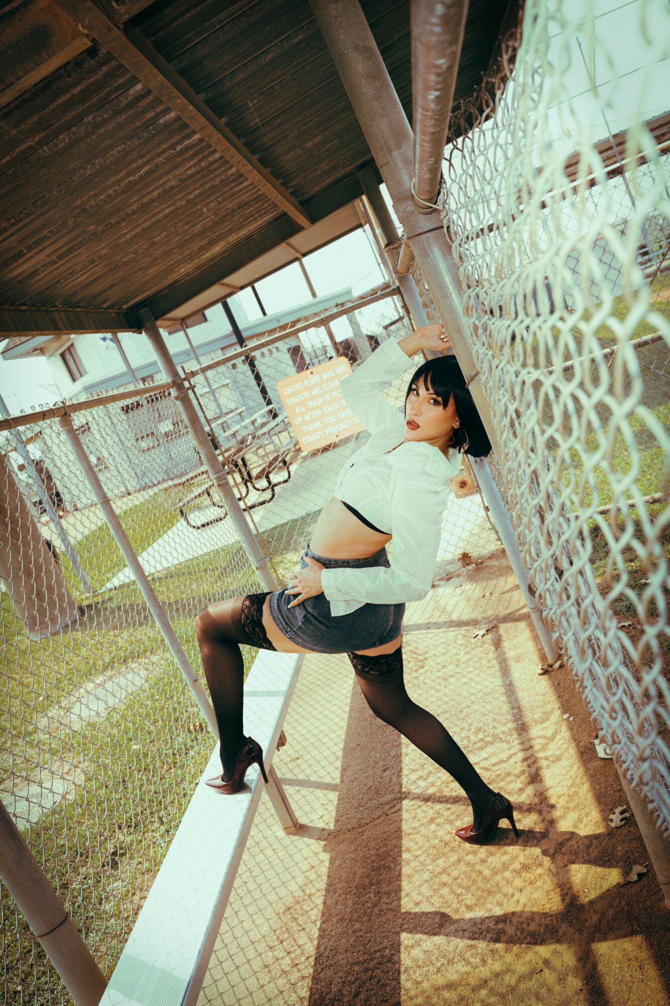 Young woman leaning against a chain-link fence under a wooden structure, with a playground in the background.