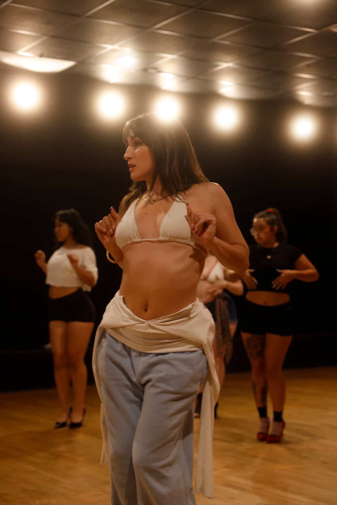Woman in a dance studio with three women in the background, wearing casual clothing, illuminated by ceiling lights.