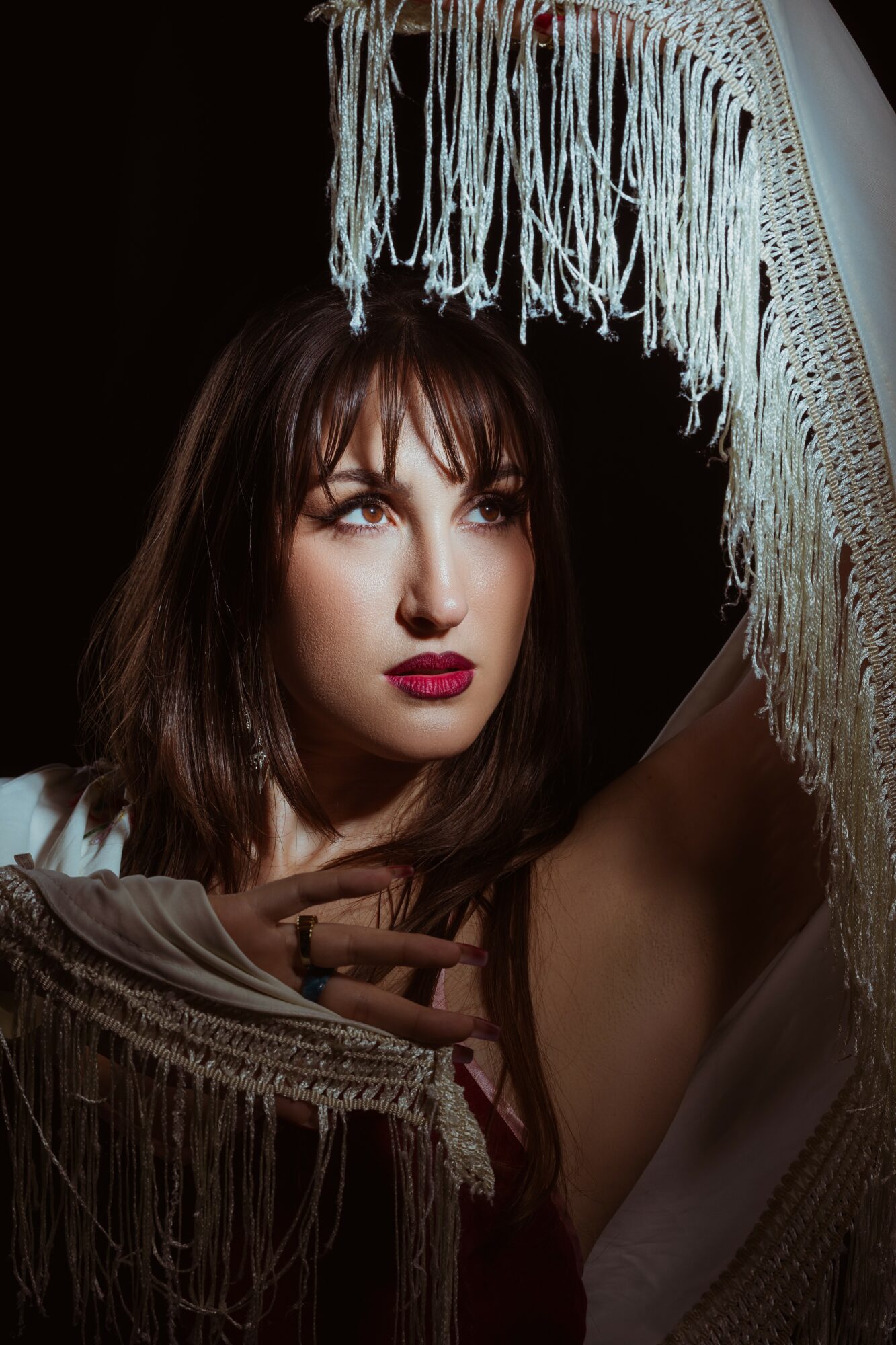 Woman with dark hair and red lipstick holding a fringed fabric, looking to the side, dark background.