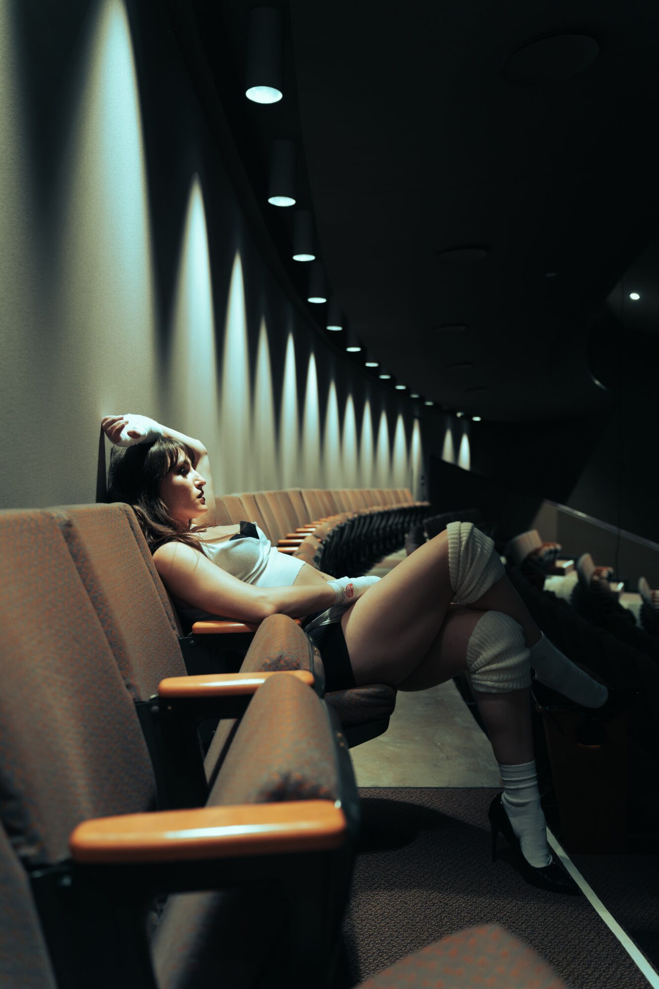 Woman reclining on theater seat with legs up, illuminated by overhead lights in dark room.