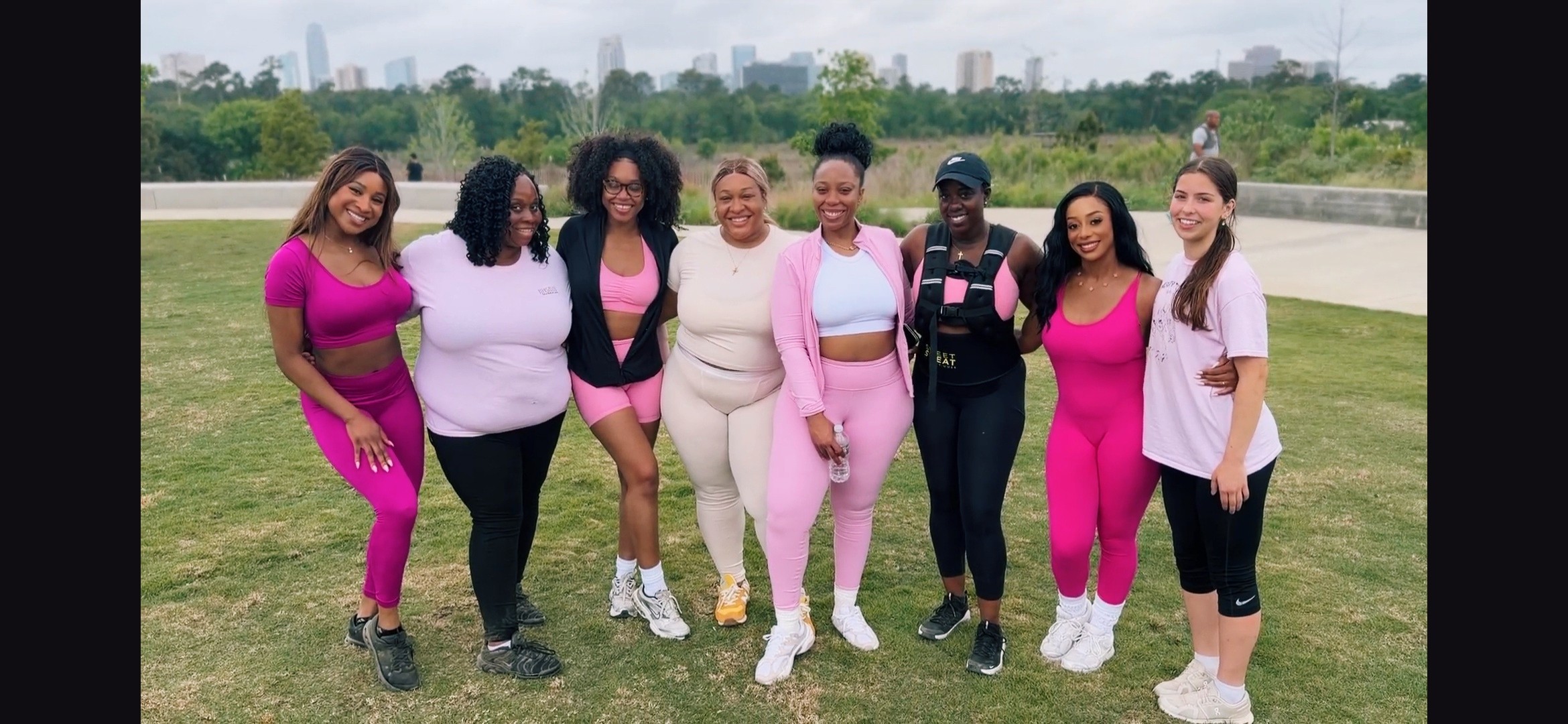 Group of eight women standing outdoors on grass, with a city skyline in the background.