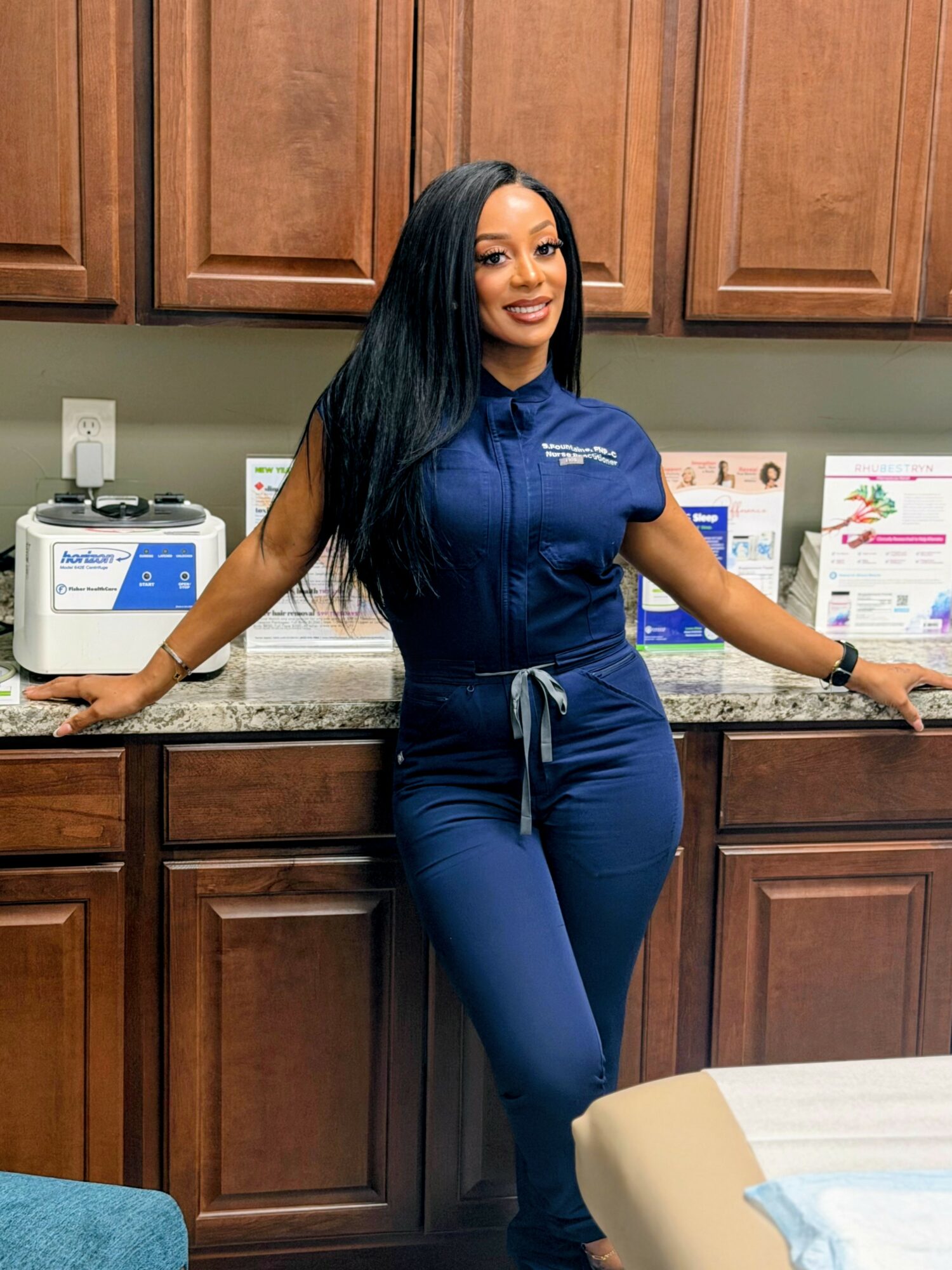 Woman in blue scrubs standing in a medical office, smiling, with wooden cabinets behind her.