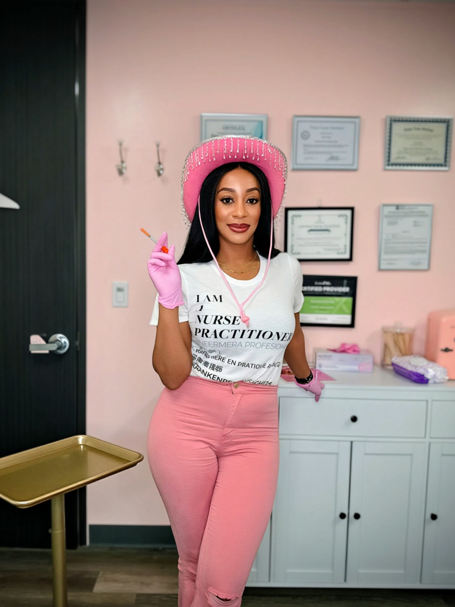Woman in pink hat and gloves smiling, standing in a room with certificates on pink wall, white cabinet, and gold tray.