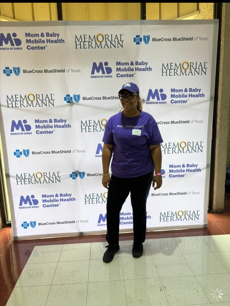 Person standing in front of a step-and-repeat banner with logos and text, wearing a purple shirt and black pants.