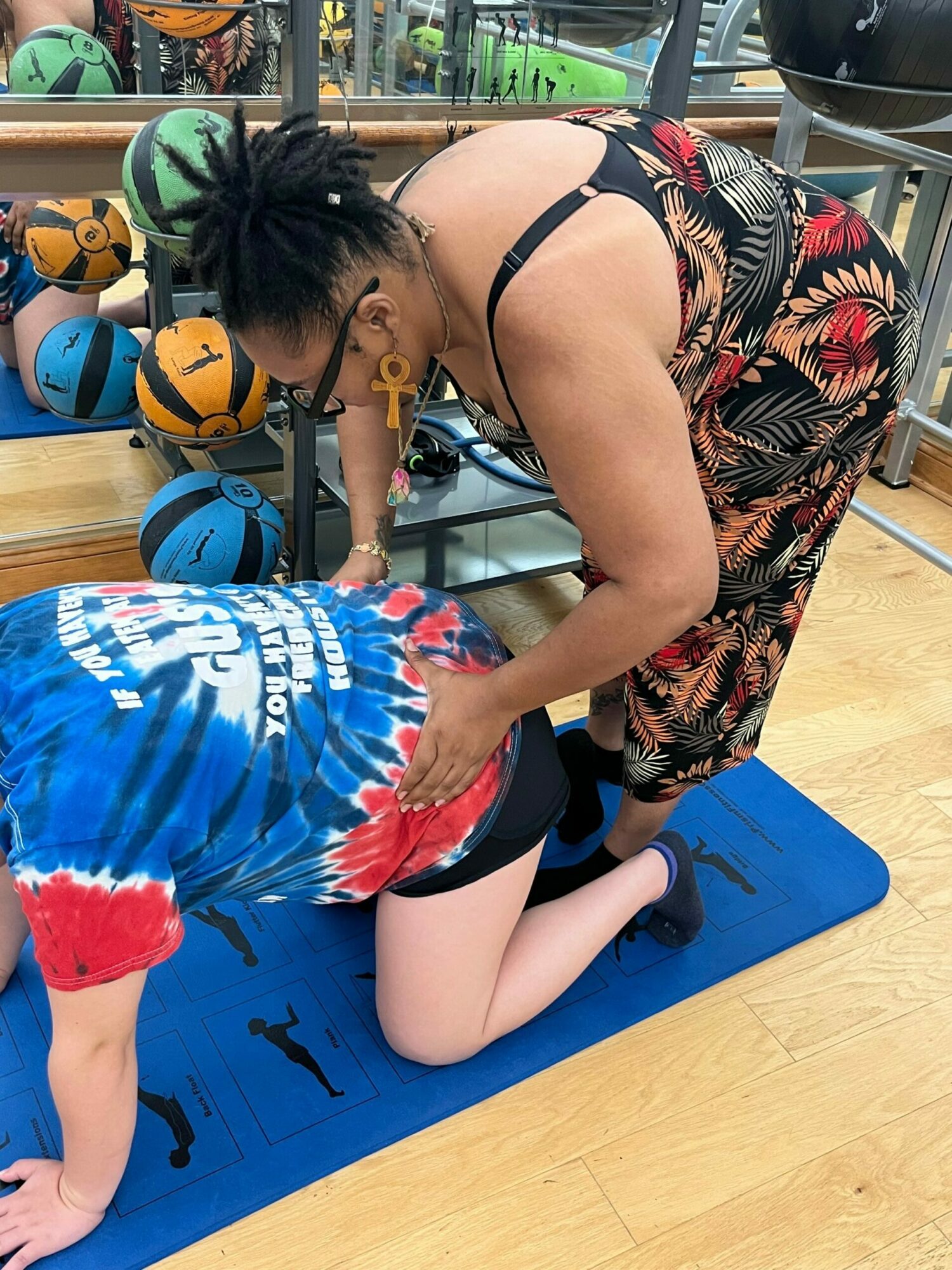 Two children, one in a tie-dye shirt and the other in a swimsuit, are on a blue mat with helmets and a volleyball nearby.