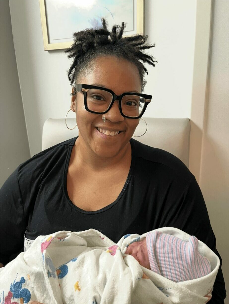 Woman with glasses smiling holding a newborn baby wrapped in a blanket, in a room with a framed picture on the wall.