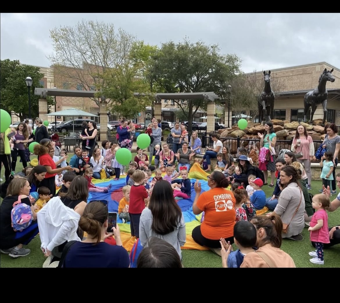 Crowd of children and adults gathered outdoors around a small pool with green balloons, trees, and buildings in background.