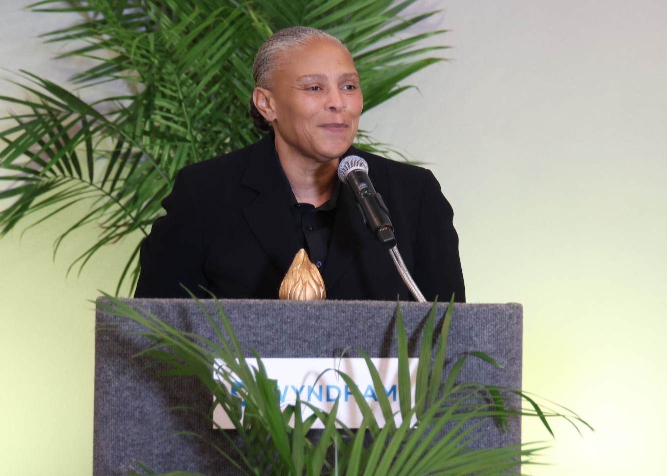 Woman speaking at a podium with a microphone, plants in the background.