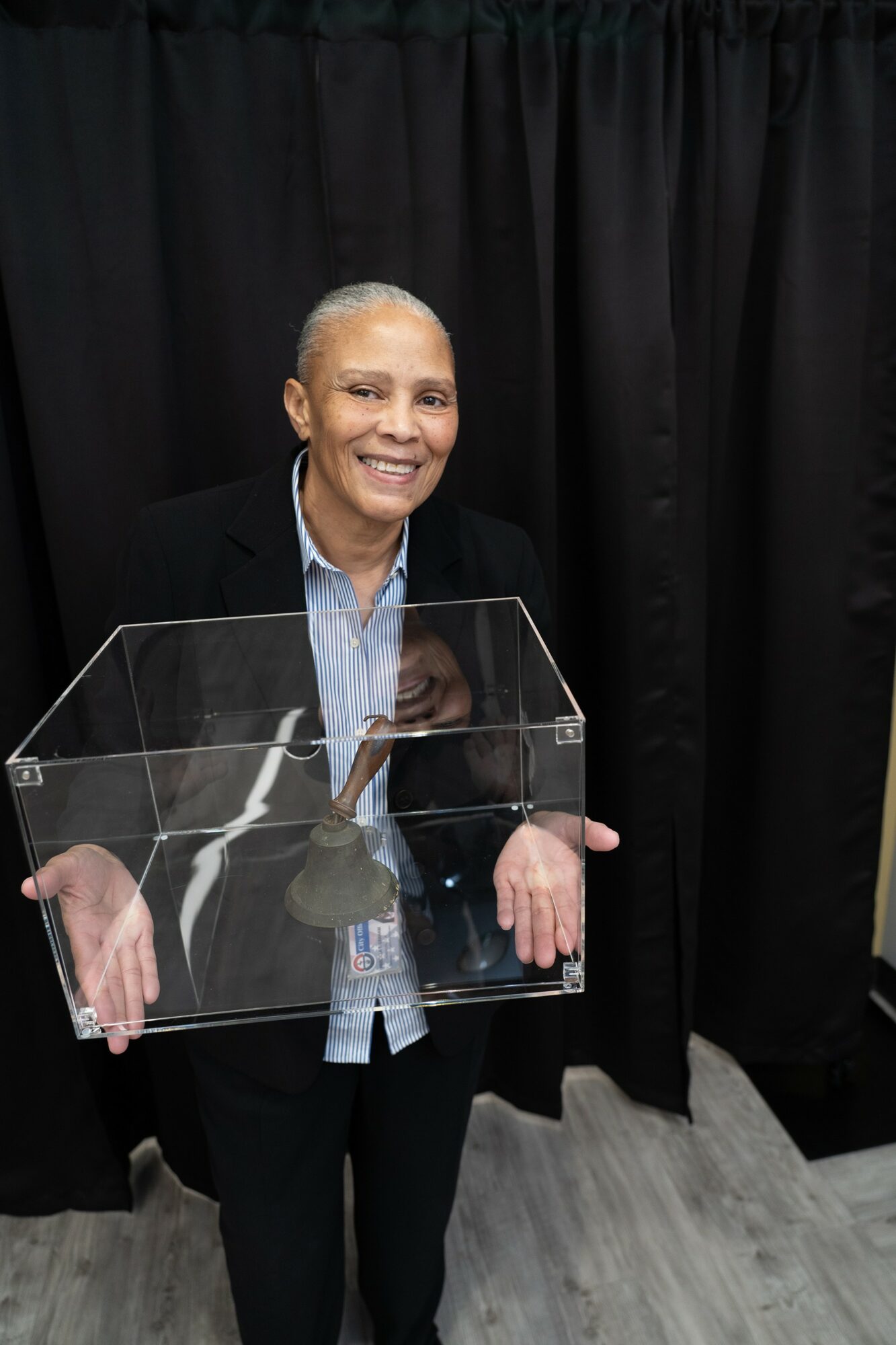 Woman smiling, holding a transparent box, standing in front of black curtains, wearing a dark blazer and light blue shirt.