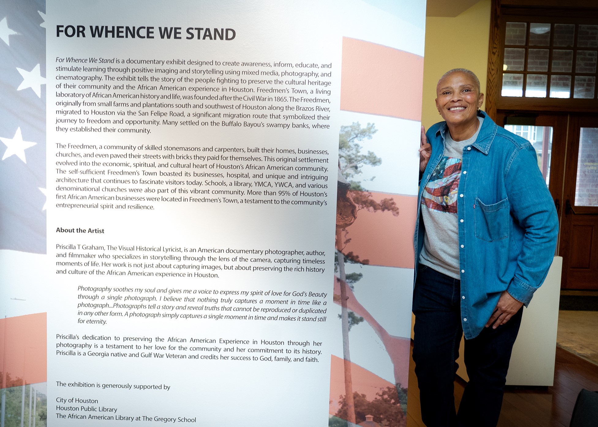 A smiling woman stands indoors near a display with a flag and text about a movement, wearing a blue jacket and jeans.