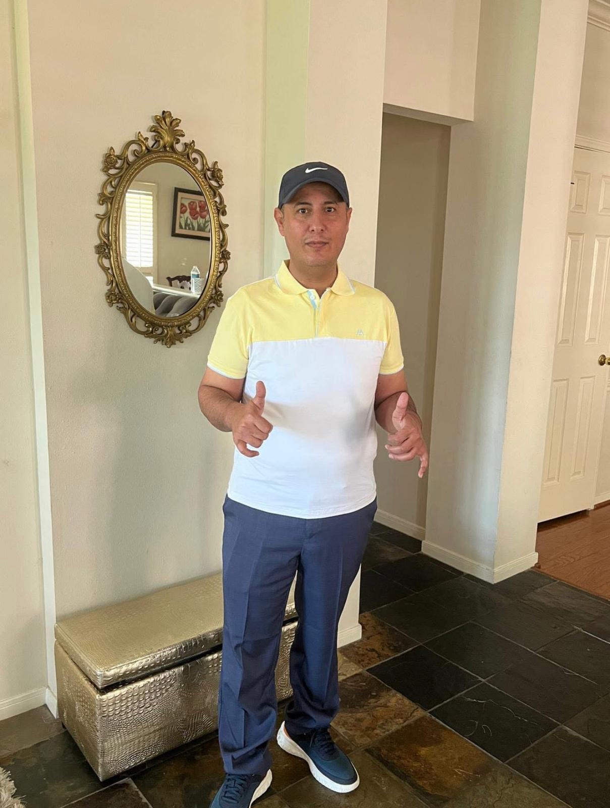 Man in yellow and white polo shirt, blue pants, and cap standing indoors, giving thumbs-up gesture.