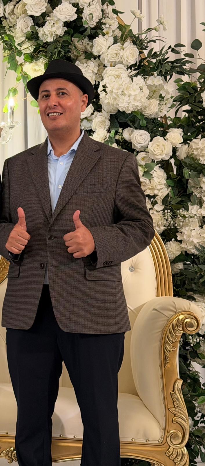 Man in a suit and hat giving two thumbs up, standing in front of a floral backdrop.
