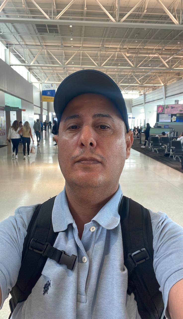 Man wearing a blue cap and light blue polo shirt with a backpack in an airport terminal.