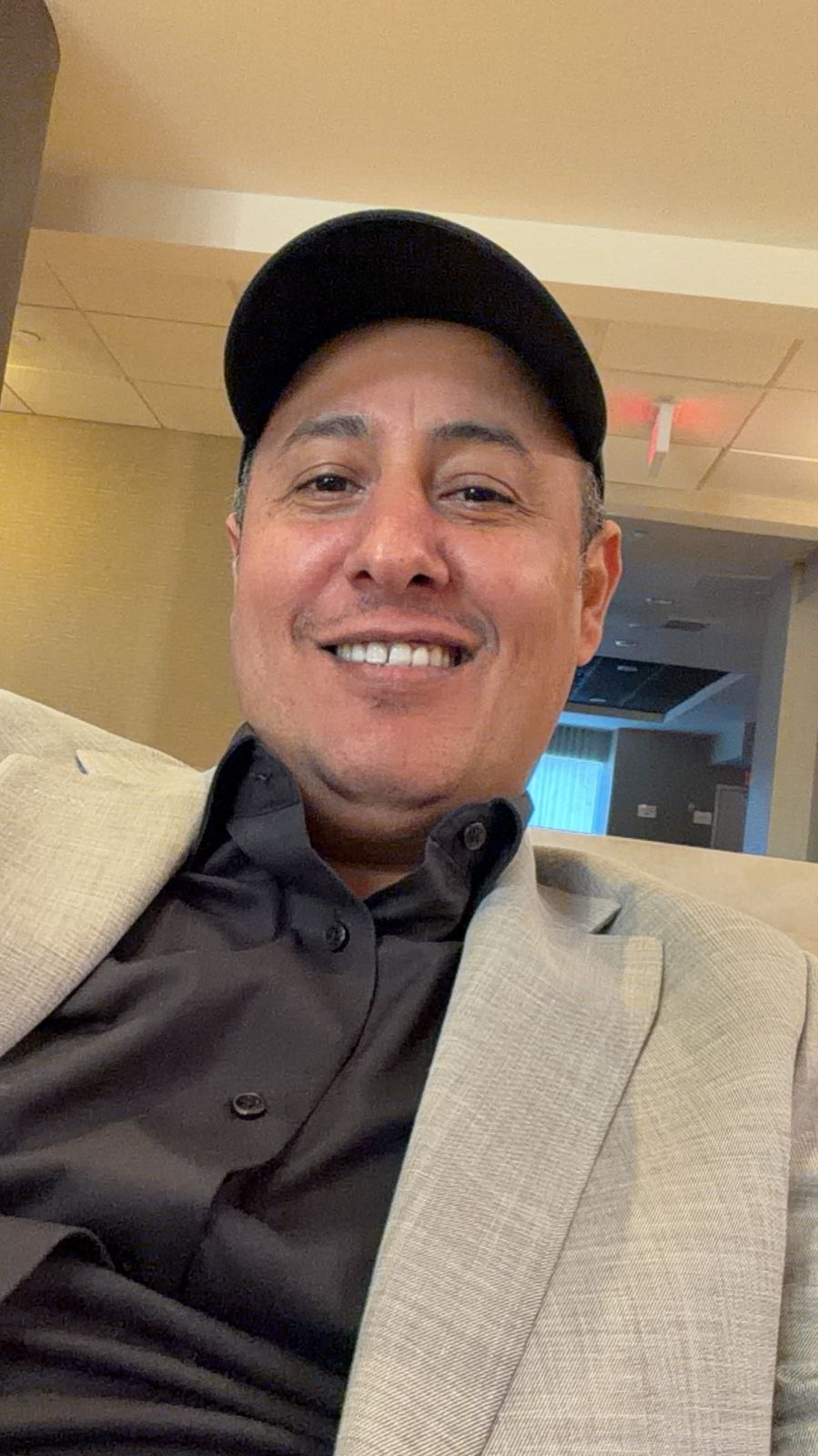 Man smiling, wearing a black cap, black shirt, and light-colored blazer indoors.