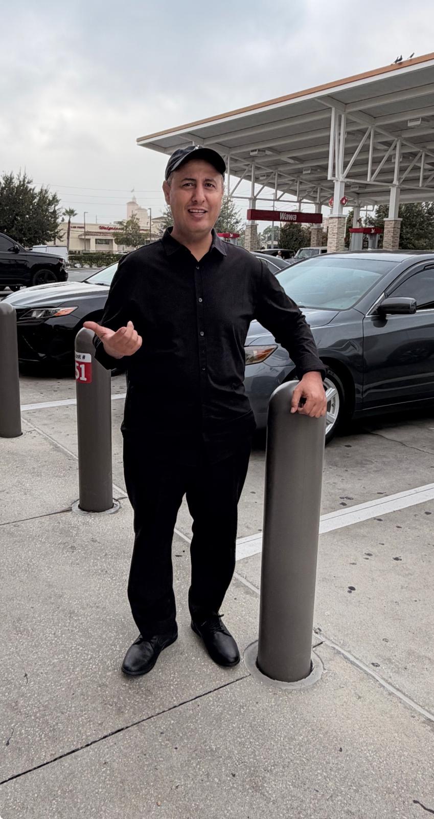 Man standing outdoors near parked cars, smiling, wearing black clothing and a cap, with a building in the background.