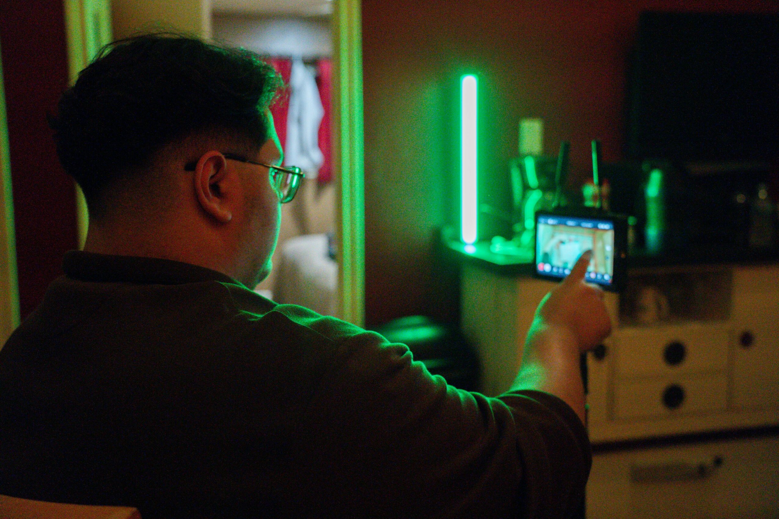 Person with glasses taking a photo with a smartphone in a room illuminated by green neon lights, facing away from the camera.