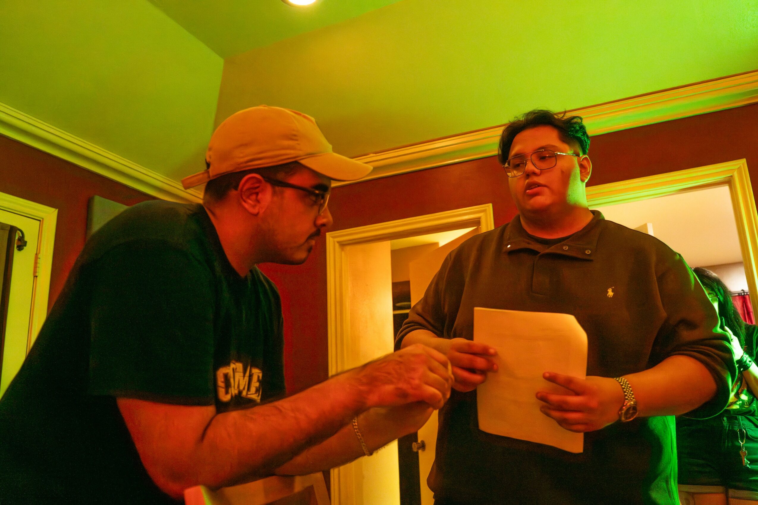 Two men standing indoors, one holding a paper, engaged in conversation, with colorful lighting and framed pictures in the background.