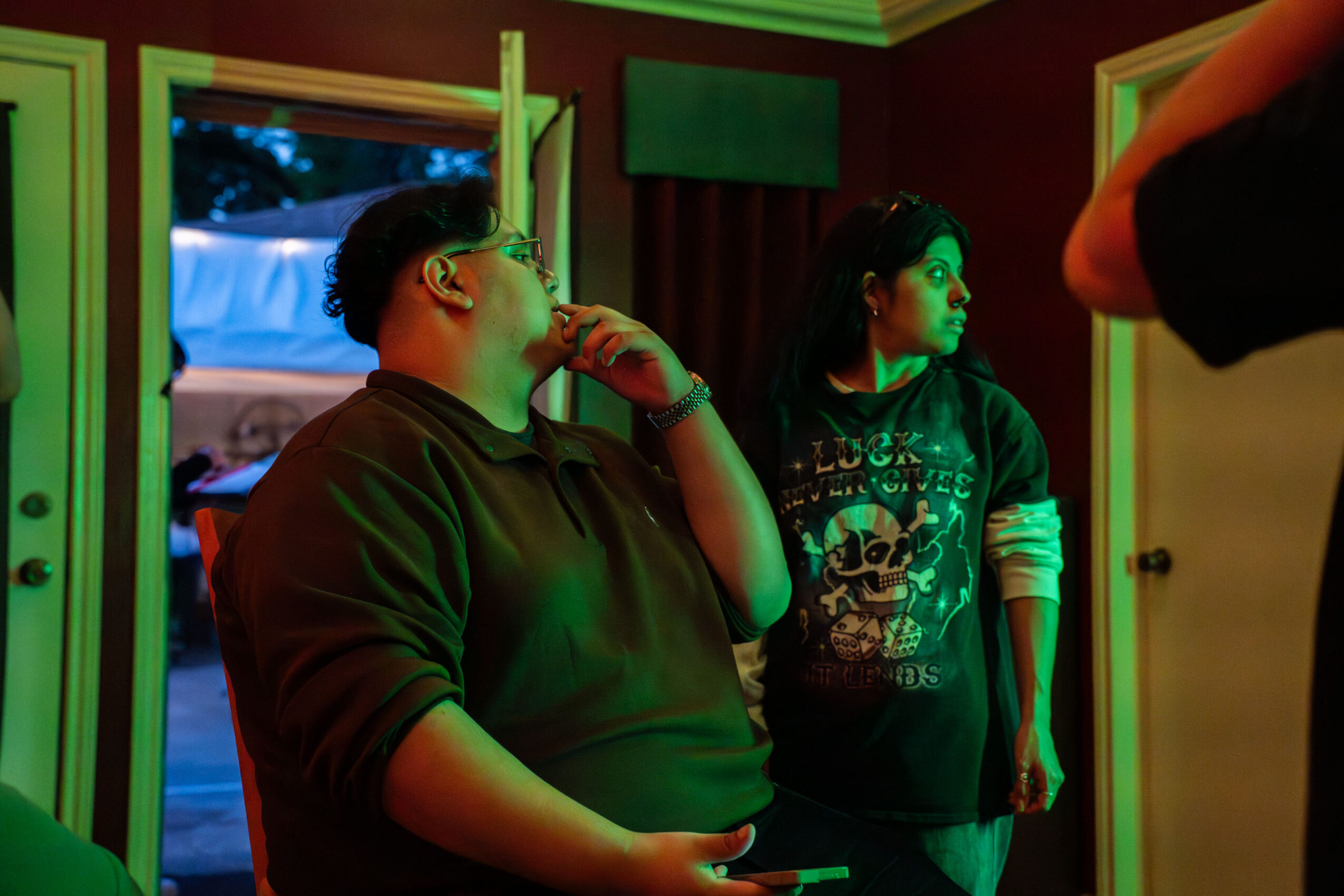 Two people inside a room with open doors, one wearing glasses and a hoodie, the other in a graphic shirt, looking to the side.