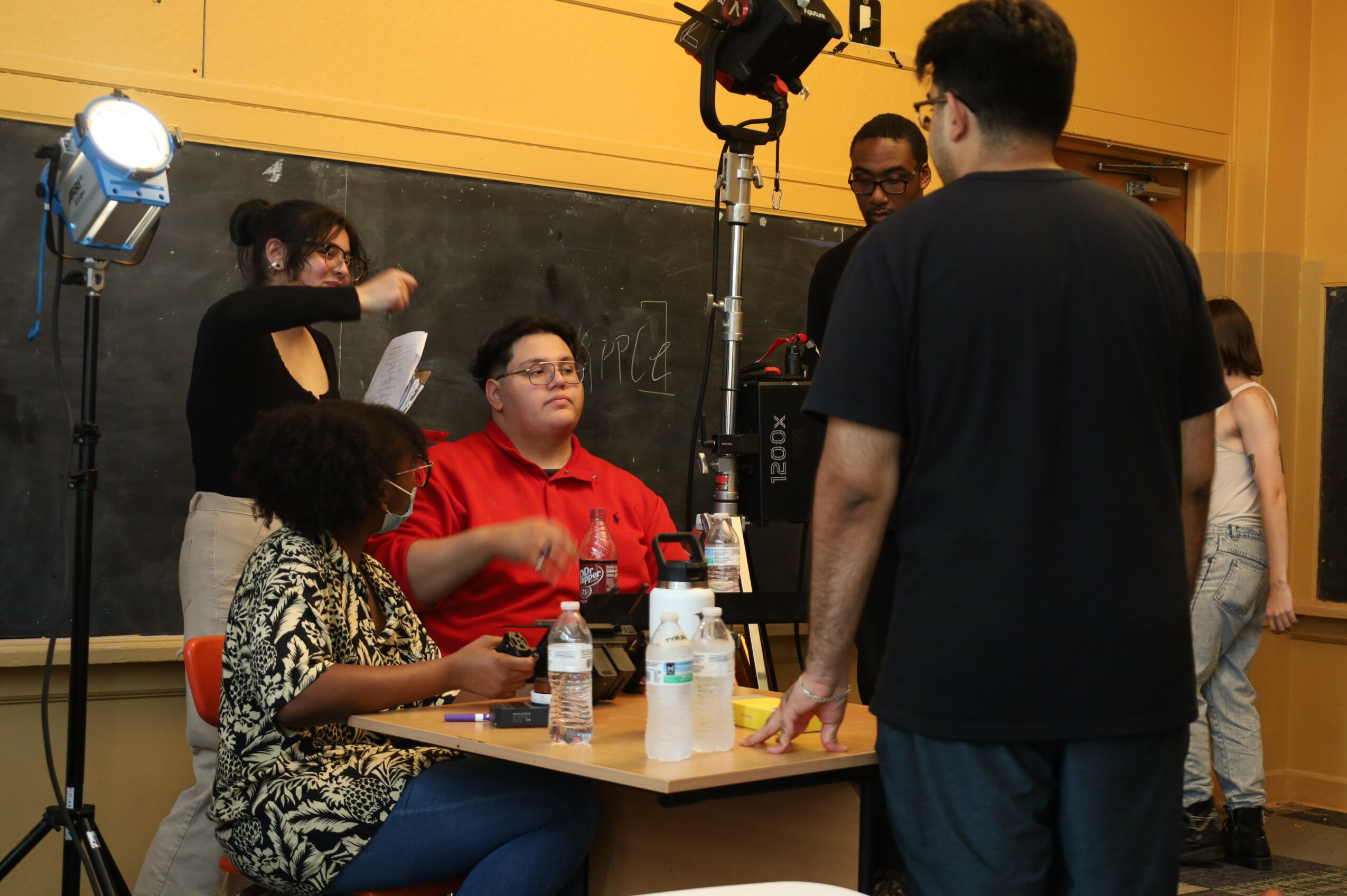 Group of people in a classroom, some sitting at a table, others standing, with filming equipment and a blackboard in the background.