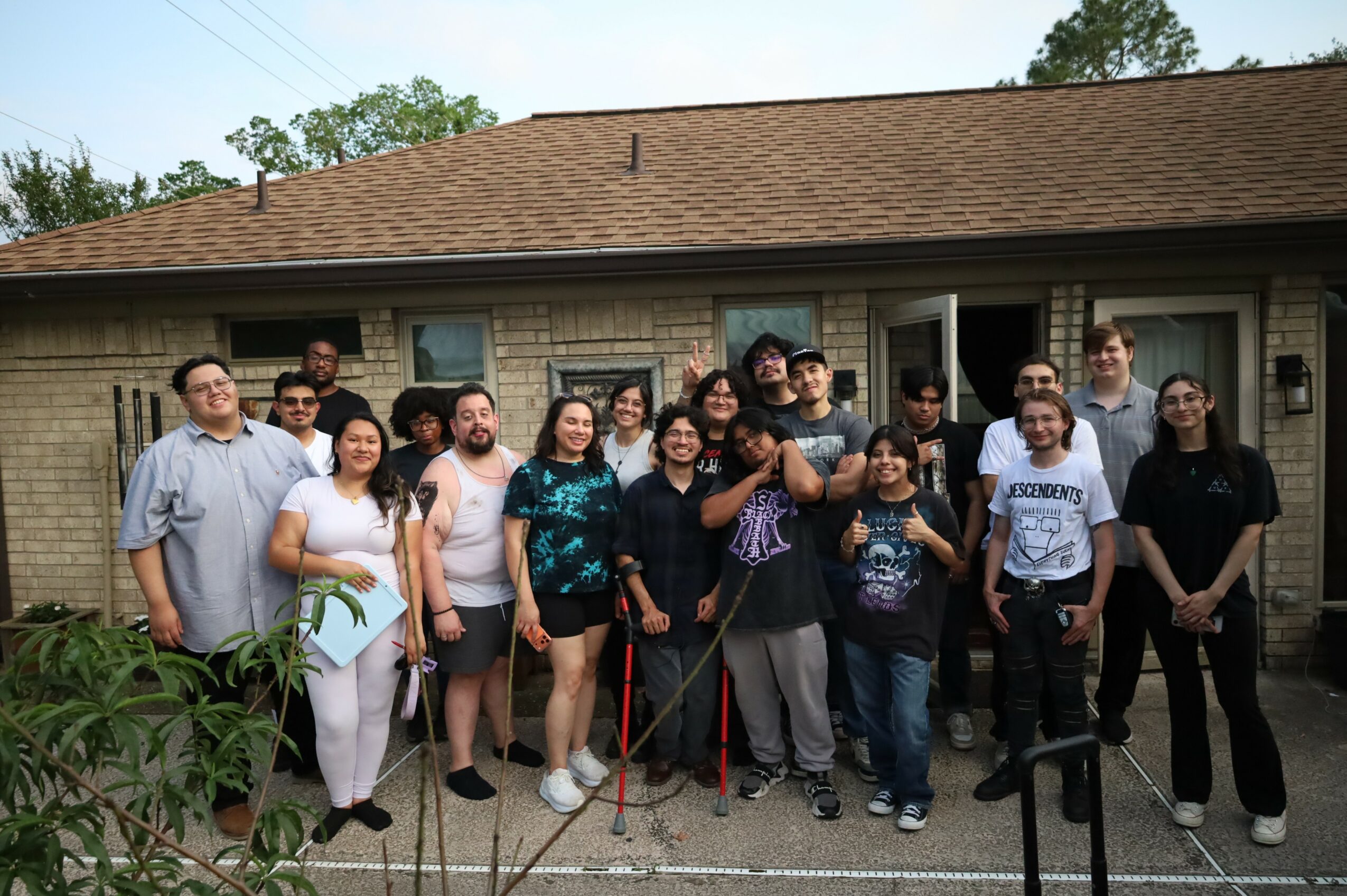 Group of people standing outside a brick building, some holding walking sticks, smiling, with trees in the background.
