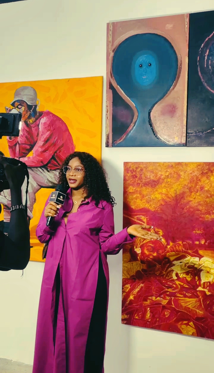 Woman in pink coat holding a microphone, standing in front of colorful abstract paintings at an art gallery.
