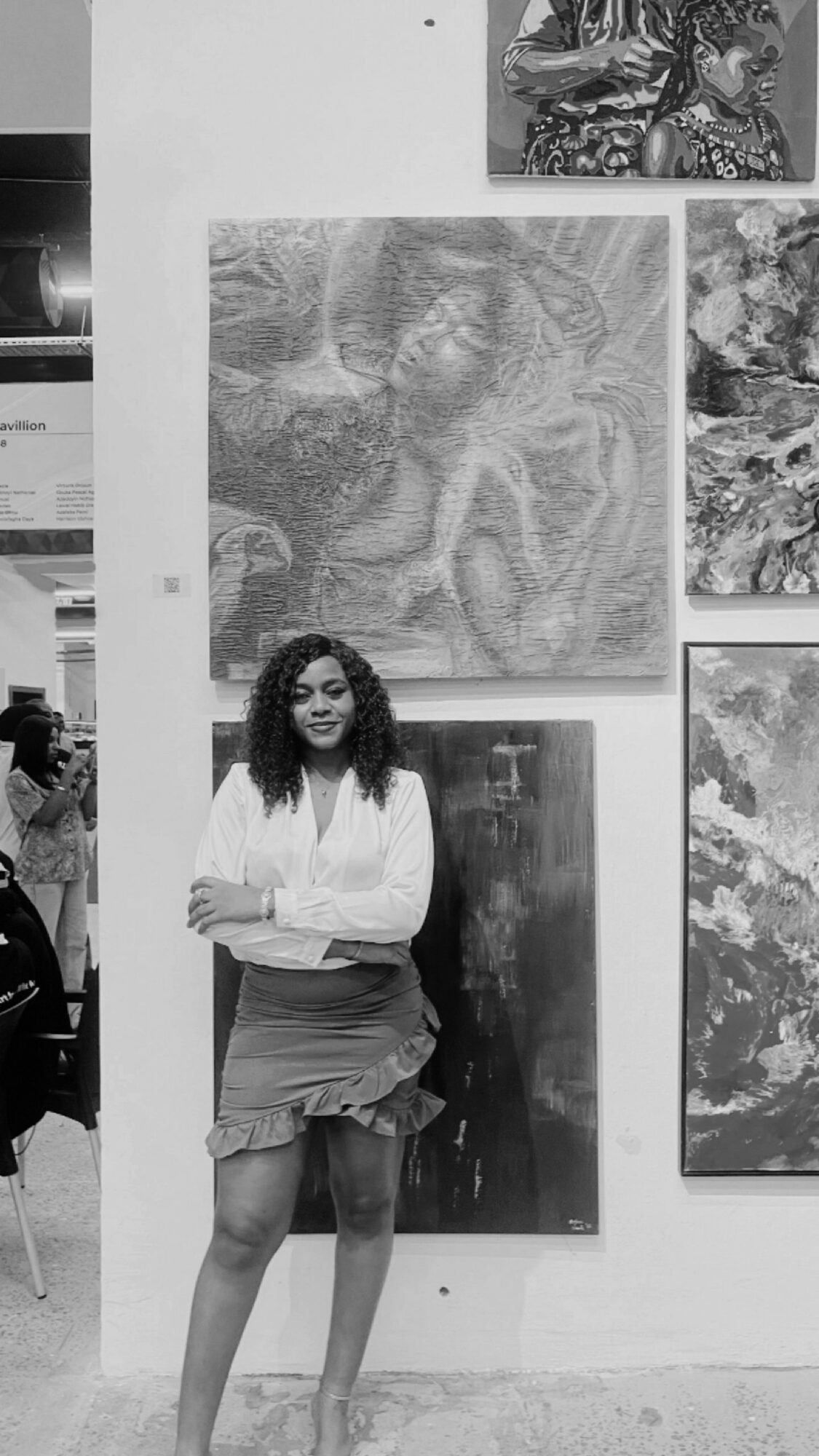 Woman with curly hair standing in front of artwork on gallery wall, smiling, arms crossed, wearing a white top and skirt.