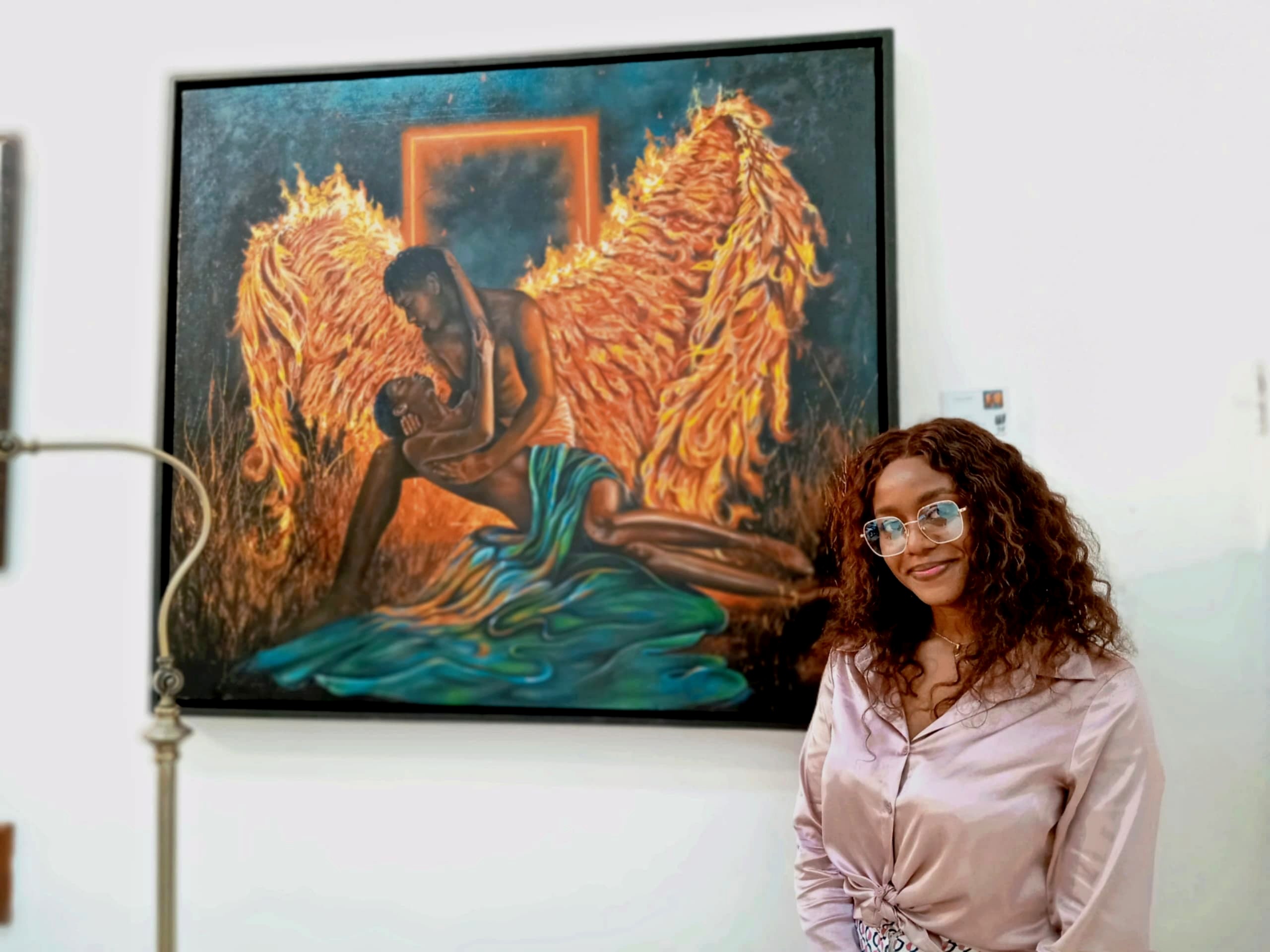 Woman with curly hair and glasses standing in front of a painting of an angel with orange wings, sitting on a blue cloth.