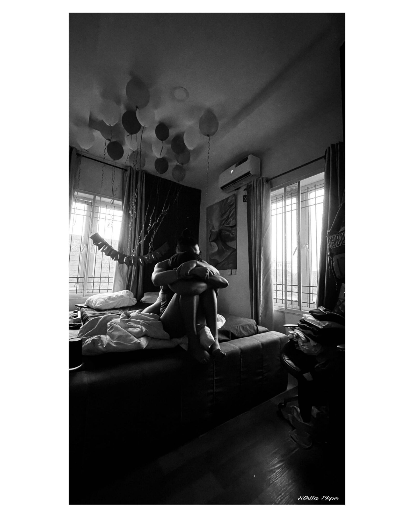 Person sitting on a bed hugging a pillow in a room with windows, curtains, and hanging ceiling lights.