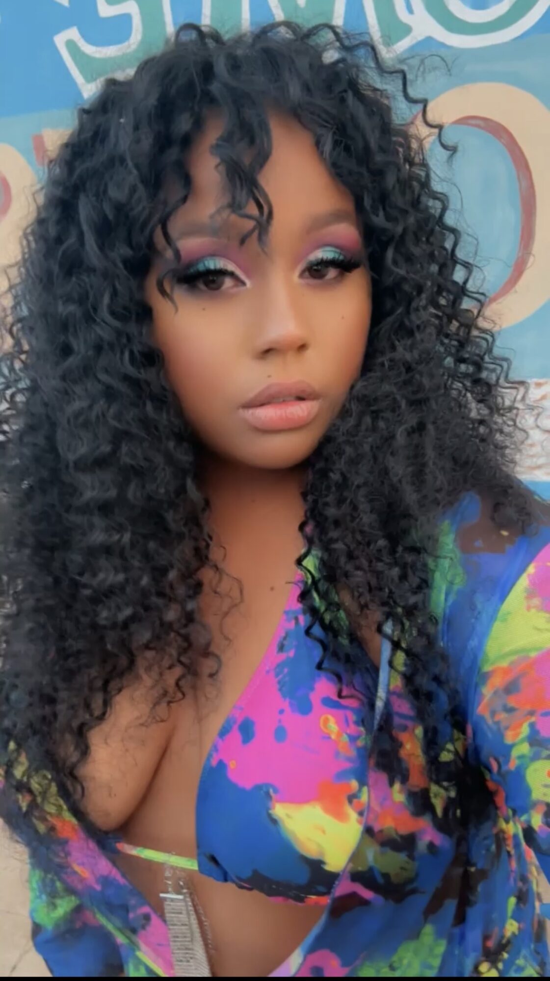 Young woman with curly hair and colorful clothing, looking at the camera with a neutral expression.
