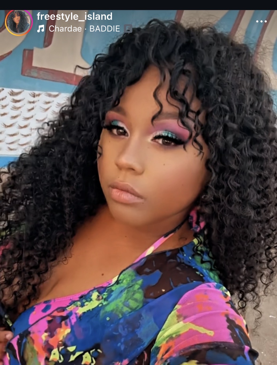 Young woman with curly hair and colorful makeup, wearing a vibrant, multicolored top, posing outdoors.