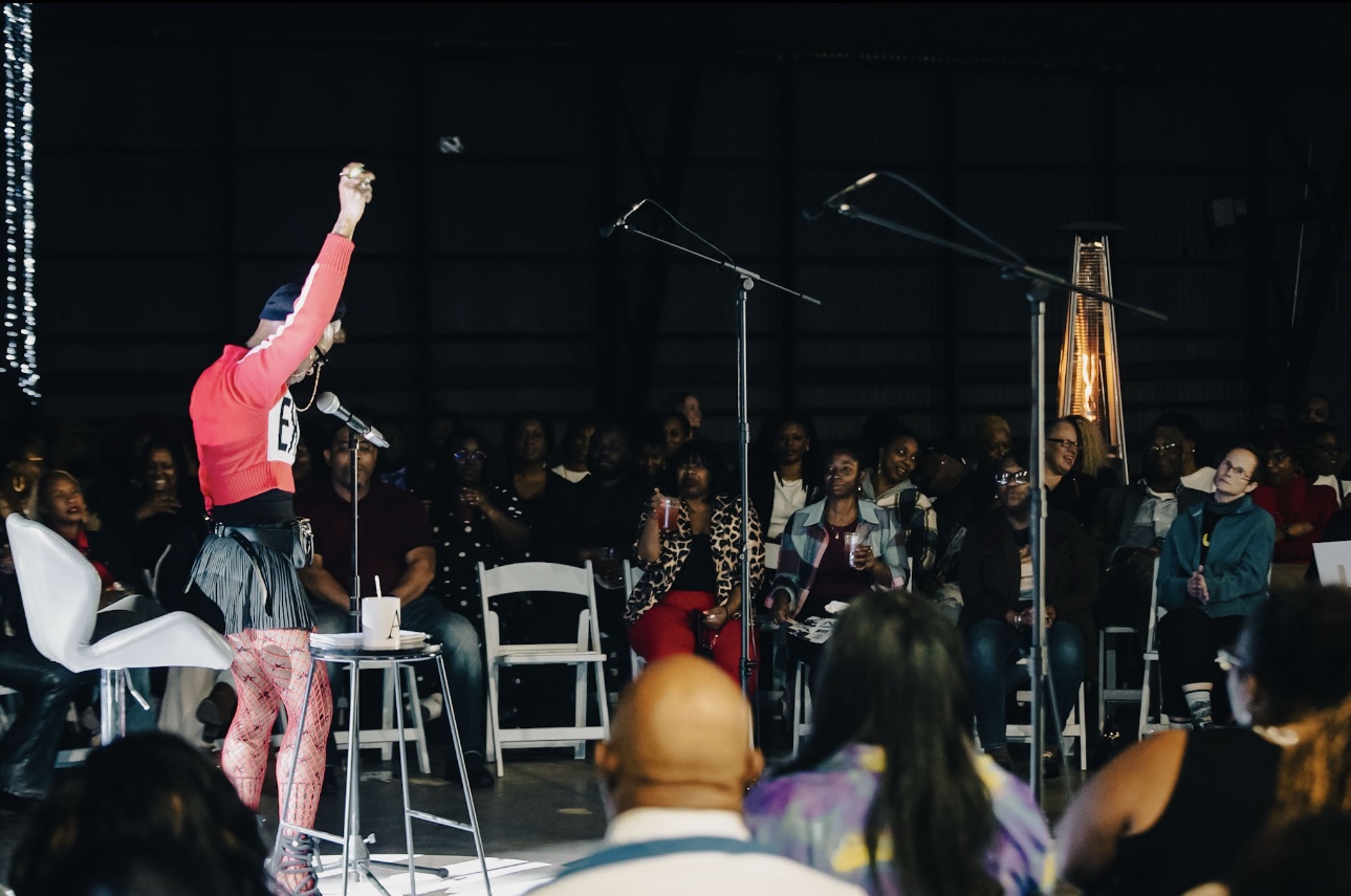 Person on stage raising an arm in front of seated audience in a dark room.